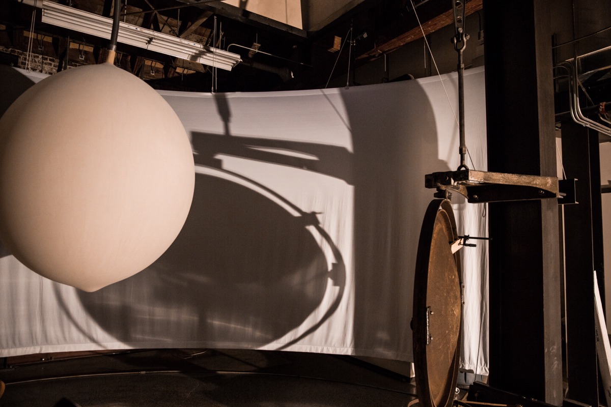 A weather balloon hangs midspan in the gallery, casting a sharp dark shadow on the screen behind it; the balloon inflated and deflated in time with Puget Sound tide data.