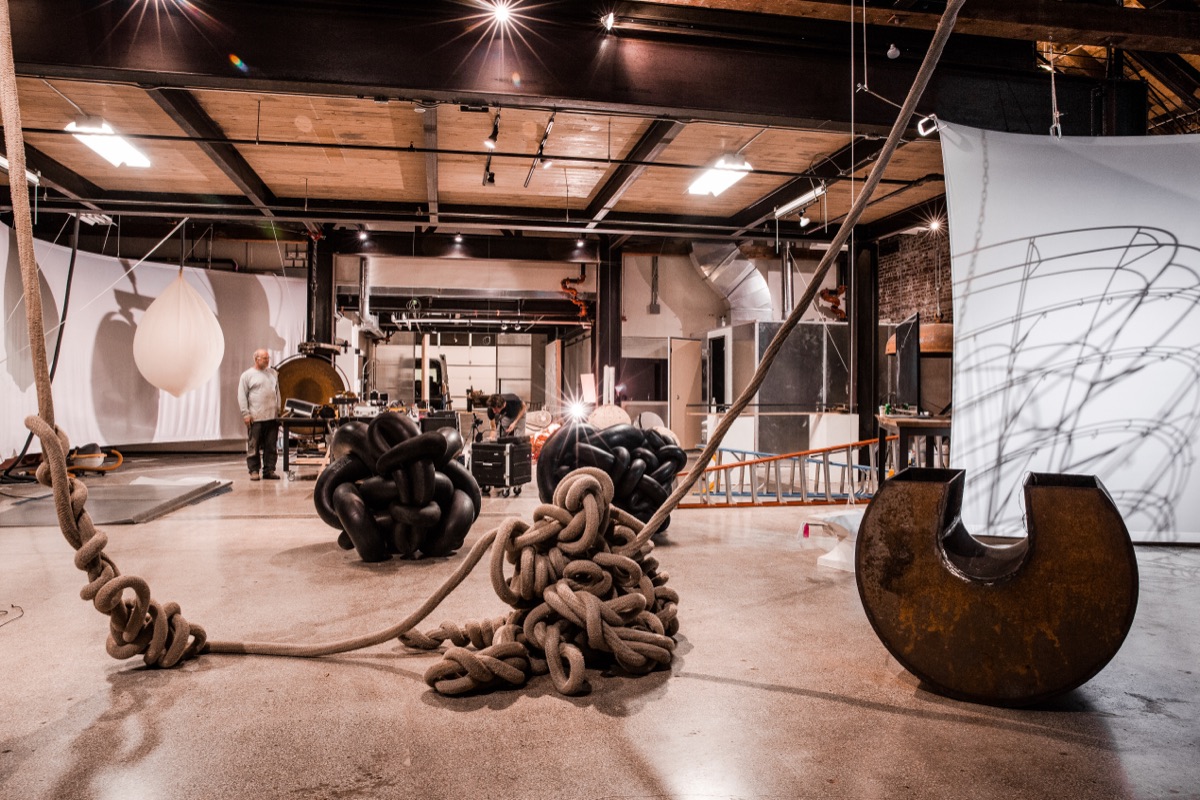 A wide gallery view: knotted rope sculptures and rusted steel hoops in the foreground, projected wire-shadow drawings on a hung screen at the rear, the bell rig visible mid-room.