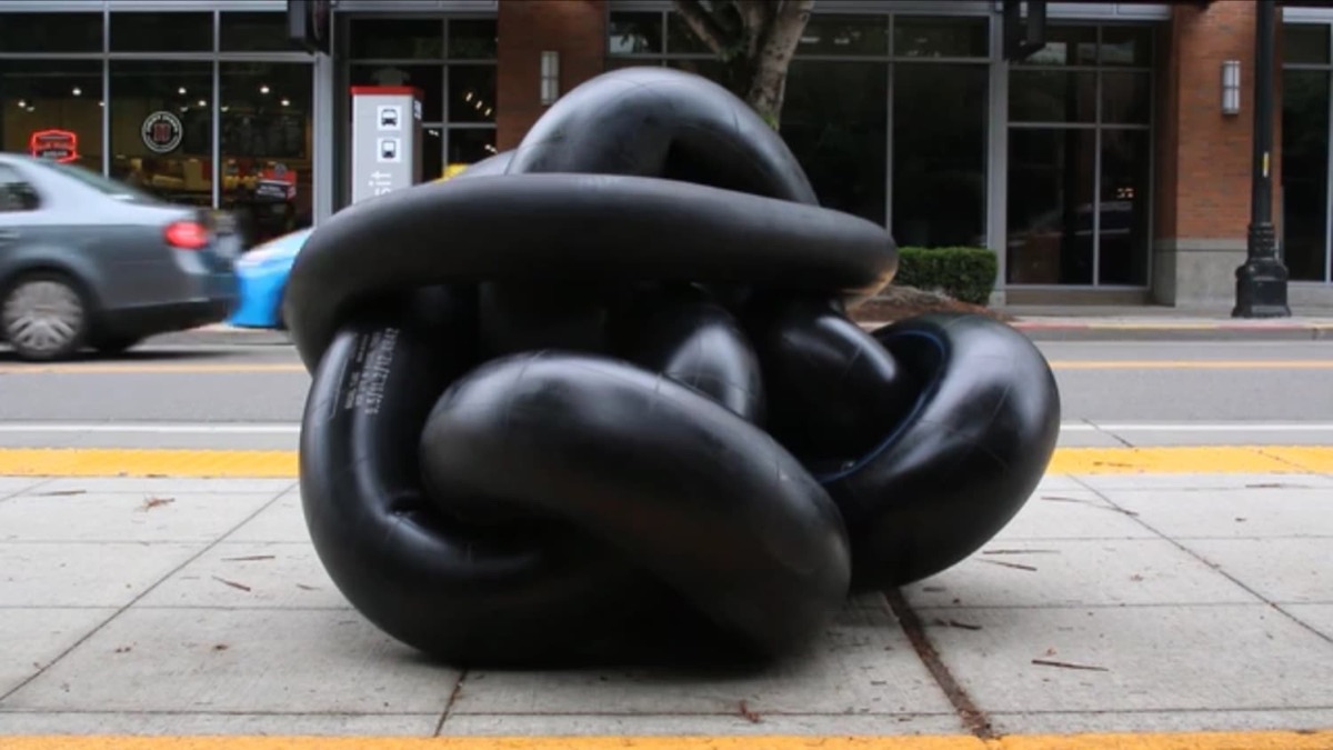 A coiled black truck-innertube sculpture sitting on the sidewalk outside the gallery, with a Seattle bus stop and a passing car in the background.