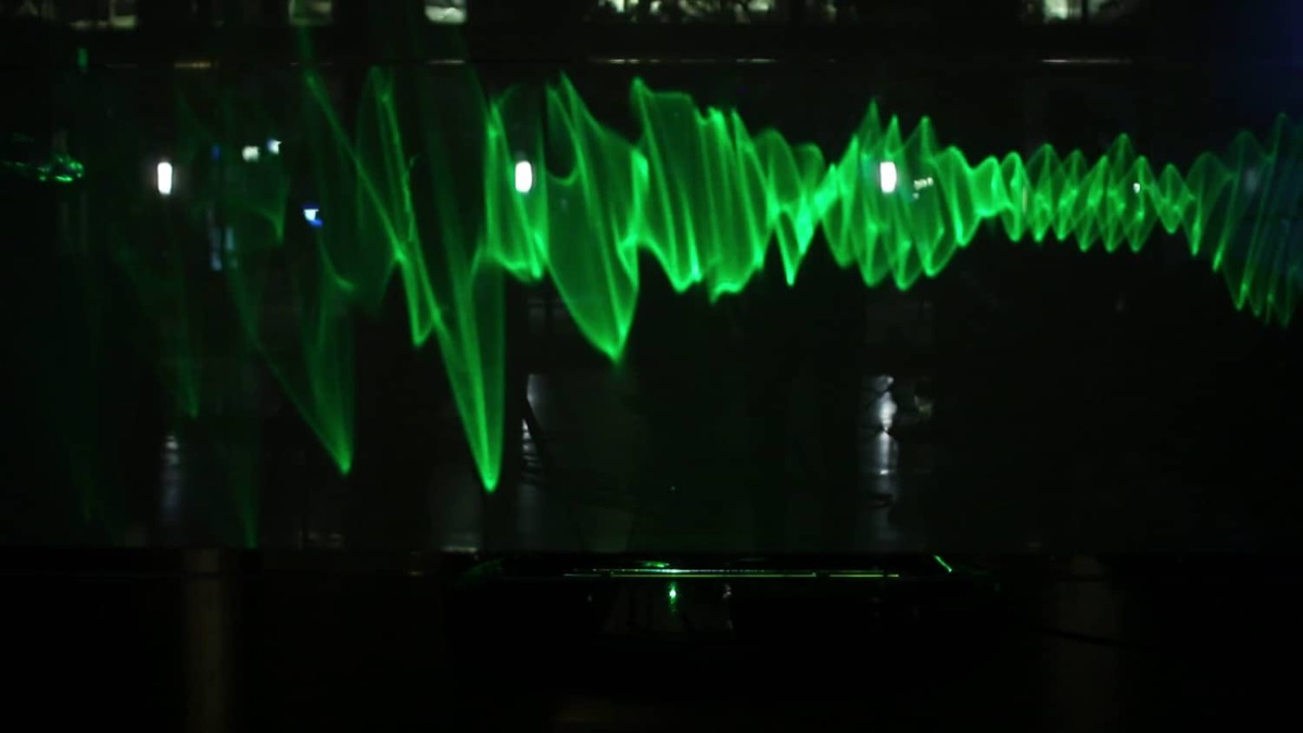 A wide green laser waveform projected across a dark backdrop — the visual signature of a bell tone passed through water.