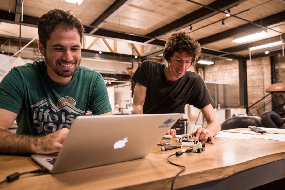 Two artists laughing over a laptop and a small electronics build at the gallery's open workbench.