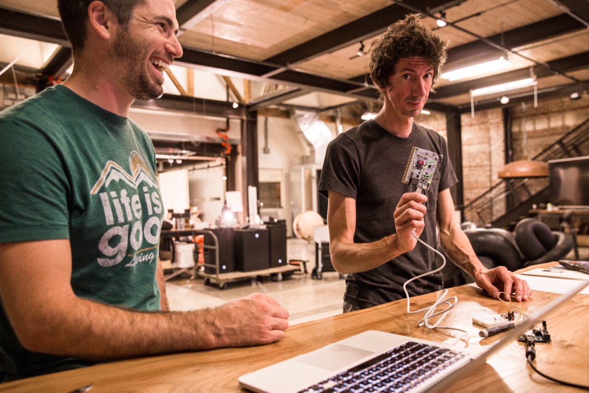 An artist holding up a small custom circuit board to show his collaborator, the gallery and tools visible behind them.