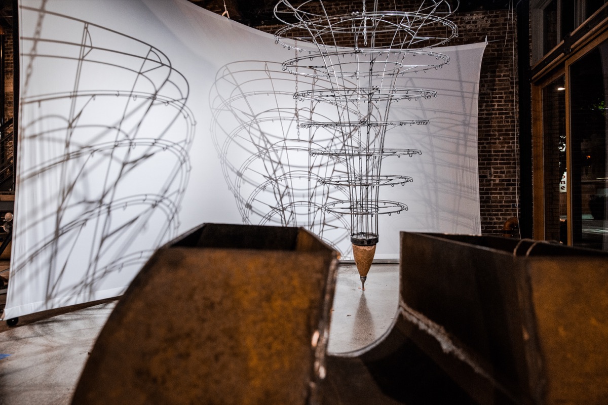 A tall conical wire-frame sculpture casting a layered shadow on a hung white screen behind it; the spiral's motion was driven by live Pacific Northwest tide data.